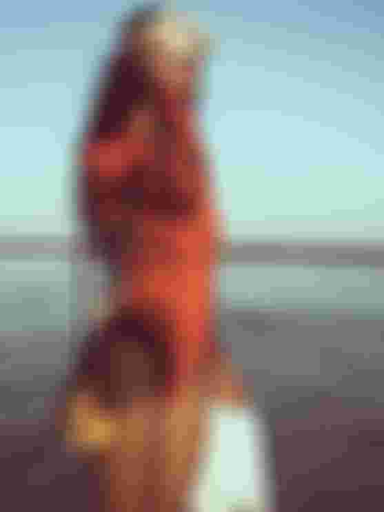 Person in orange wetsuit by the sea, holding white flippers, wearing a snorkel mask on head, with a yellow camera slung over shoulder.