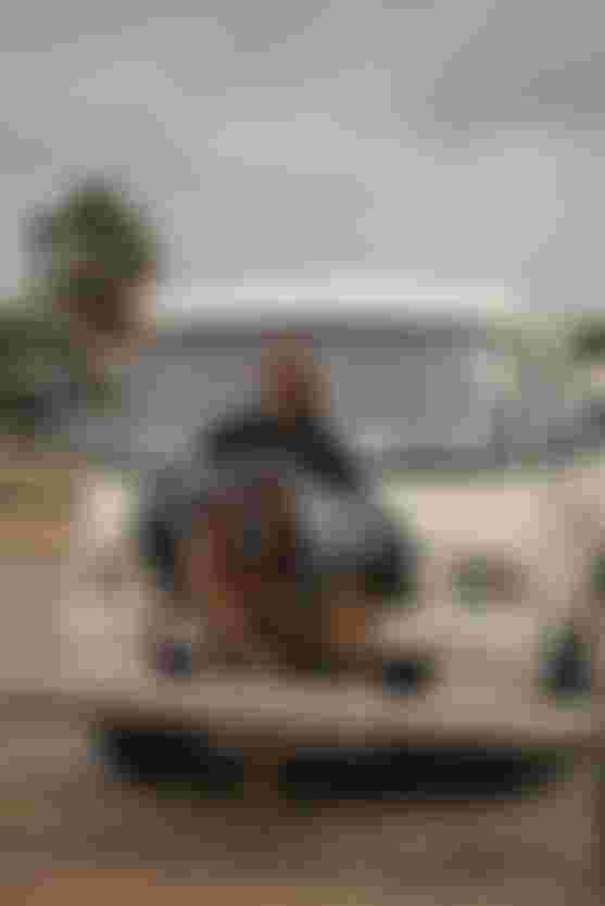 Smiling person in sunglasses stands in front of a white van holding colorful ktie harnesses, with a palm tree and overcast sky in the background.