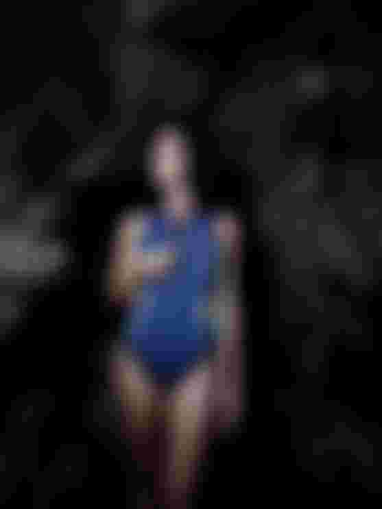 Person in a wetsuit stands in a rocky cave, looking to the side. The lighting emphasizes the texture of the rocks.
