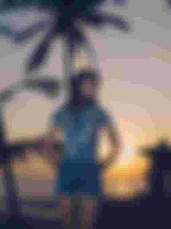 Woman in blue wetsuit stands on a beach at sunset, with palm trees silhouetted in the background.