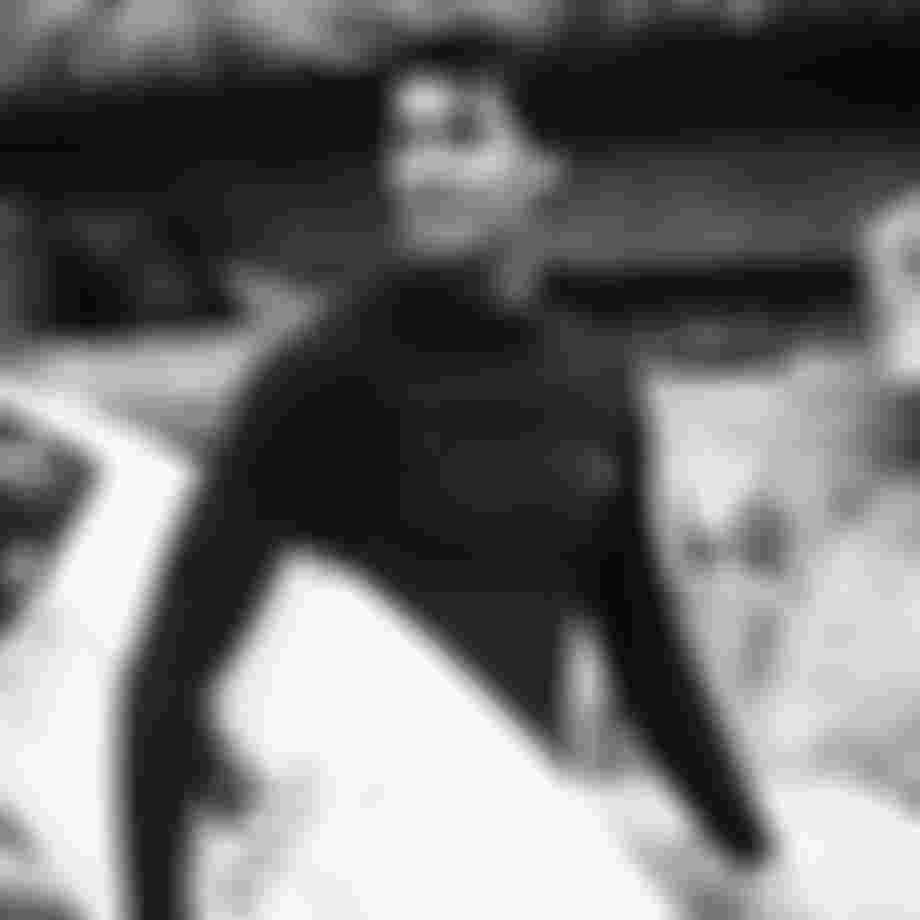Surfer in a wetsuit holding a surfboard, standing in front of rocky cliffs, looking off into the distance. Black and white photo.