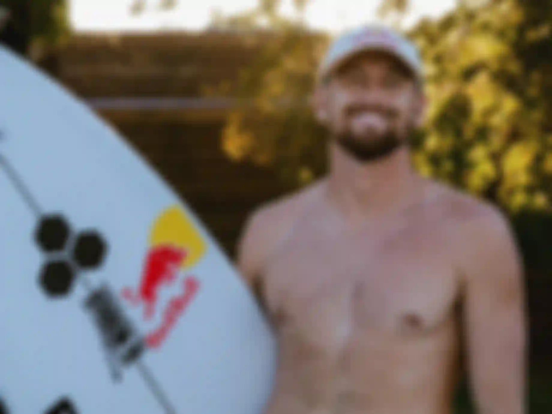 Smiling shirtless male surfer holding a Red Bull-branded surfboard outdoors in warm golden light, wearing a Red Bull cap.