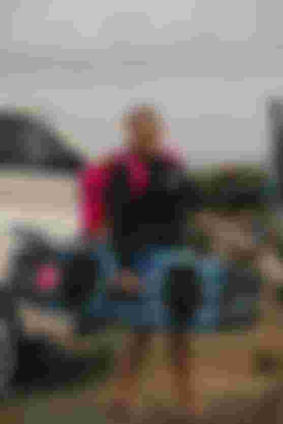 Smiling woman with a pink backpack holding a kiteboard, standing barefoot near a car and a cloudy beach scene.