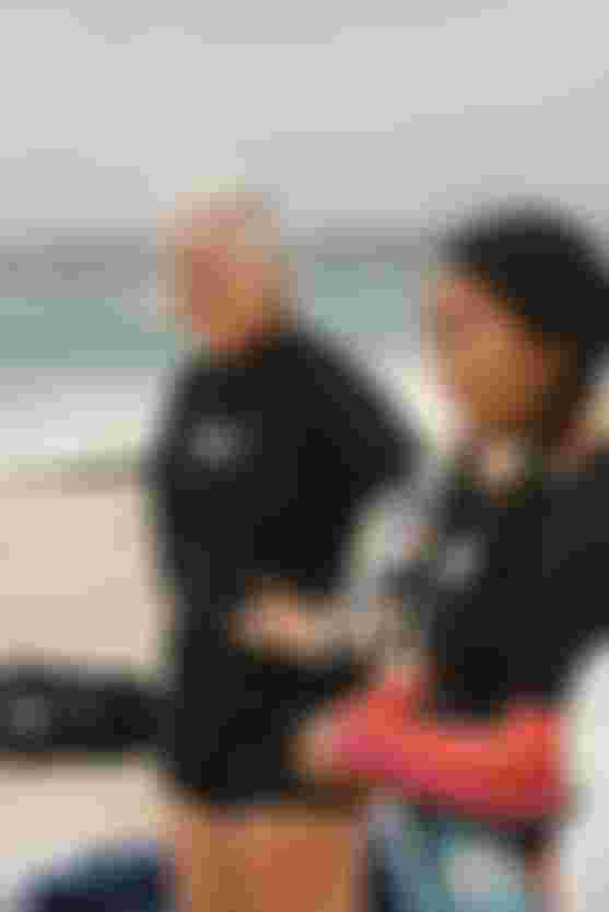 Two people in wetsuits stand on a beach, looking towards the ocean. The sky is cloudy, and surfboards are seen in the background.