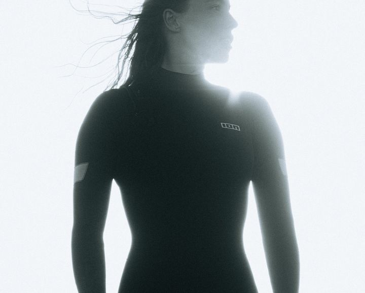 Silhouetted person in a wetsuit stands against a bright sky, with sunlight highlighting their outline and hair gently blown by the wind.