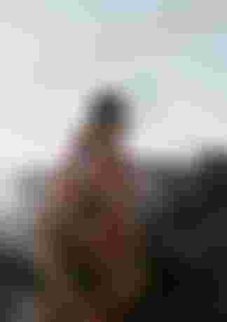 A person in a colorful hooded garment stands near large rocks by the sea, with a ghostly overlay of their face beside them.