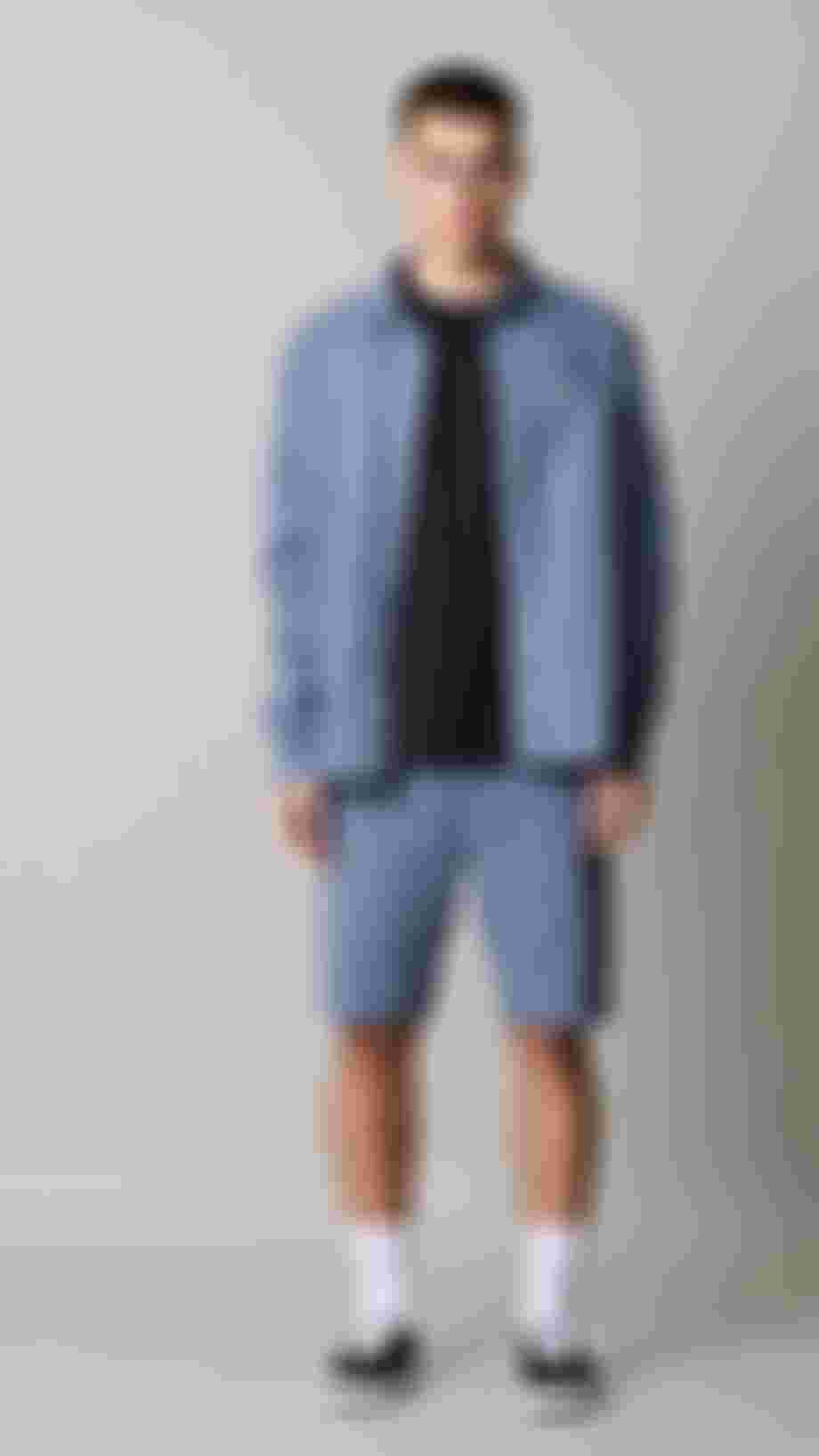 Man standing in front of light grey background wearing blue denim shorts and a loose denim shirt
