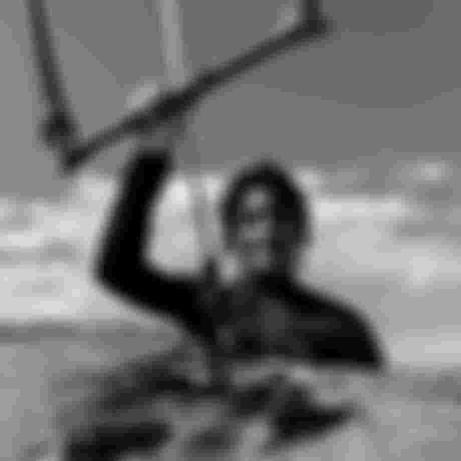 Person in a wetsuit holding a kite handle, knee-deep in water, with clouds and mountains in the background. Black and white photo.