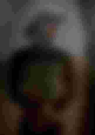 Man in a black and gray wetsuit, wearing a sun hat, stands in a thoughtful pose against a blurred, sandy background.