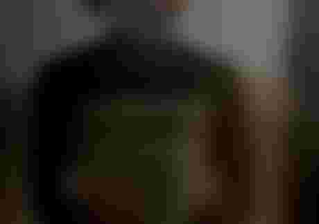 Man in a black and gray wetsuit, wearing a sun hat, stands in a thoughtful pose against a blurred, sandy background.