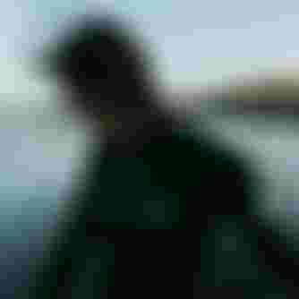 Person in a wetsuit with windblown hair stands near the ocean. The background shows gentle waves and a building in the distance.