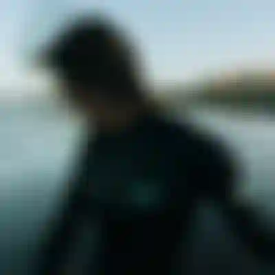 Person in a wetsuit with windblown hair stands near the ocean. The background shows gentle waves and a building in the distance.