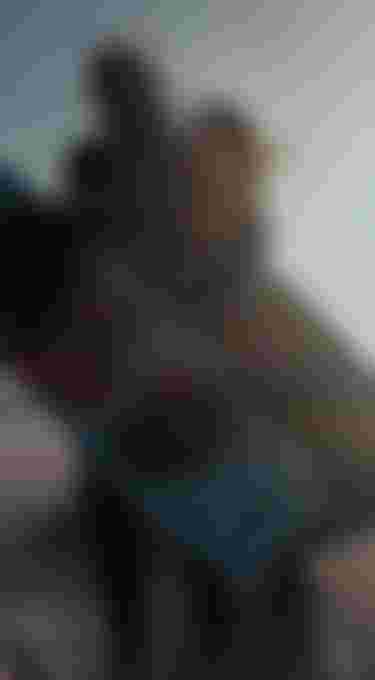 Two people in wetsuits holding colorful kitesurfing boards stand on a beach, their hair blowing in the wind, with the ocean in the background.