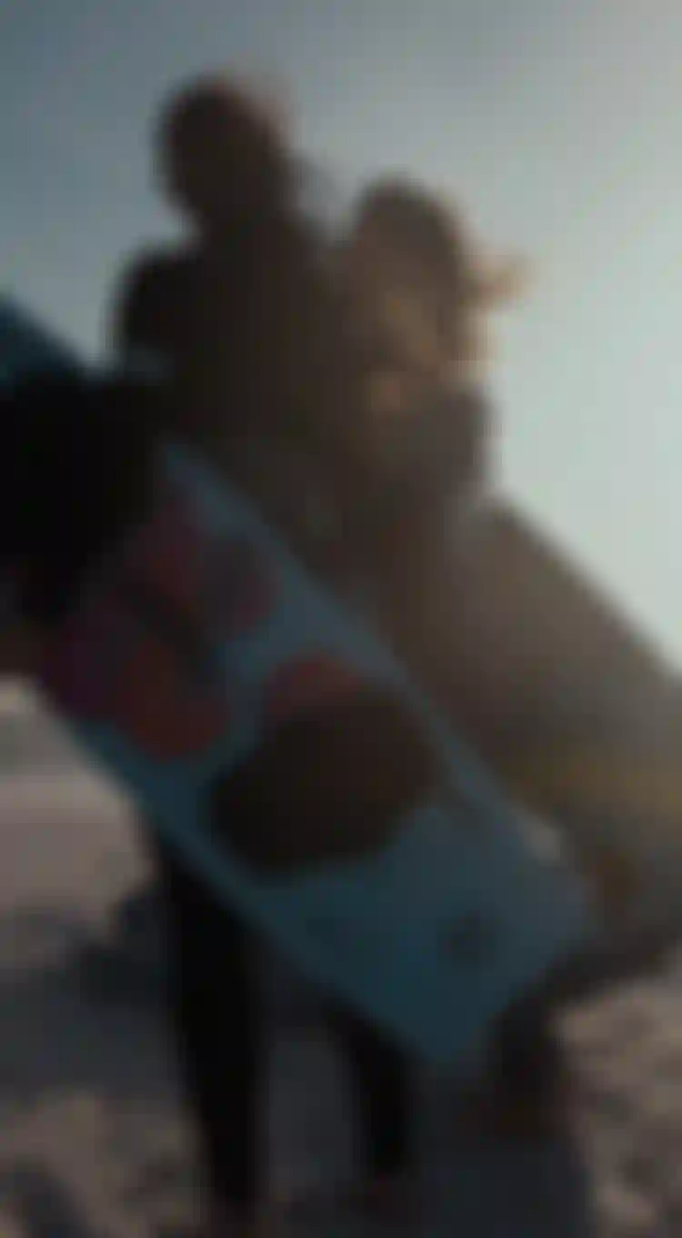 Two people in wetsuits holding colorful kitesurfing boards stand on a beach, their hair blowing in the wind, with the ocean in the background.