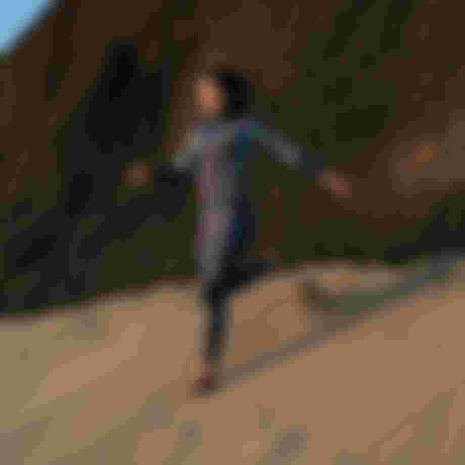 A person in a wetsuit runs barefoot on a sandy slope against a rocky backdrop, with a blue sky overhead.