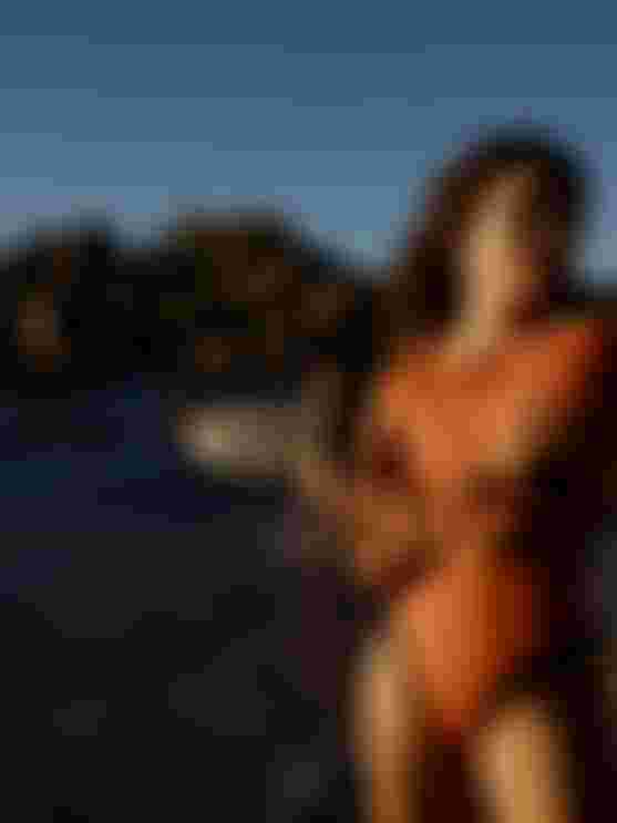 Woman in an orange wetsuit stands on a rocky beach, another woman carries a surfboard in the background under a clear blue sky.