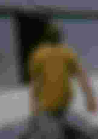 Man with long hair and a yellow T-shirt carrying surfboards towards a metal building with an open door.