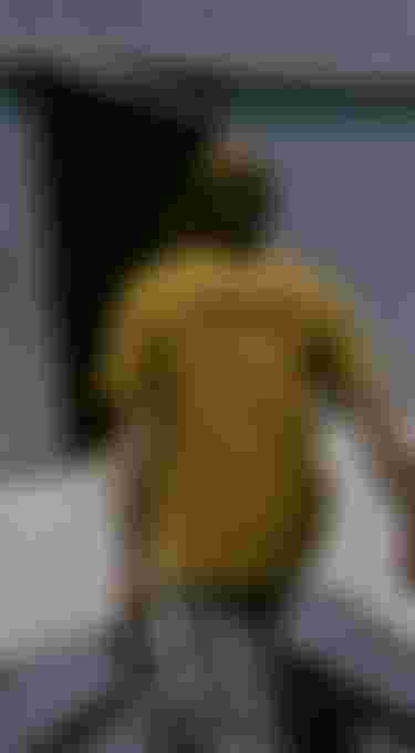 Man with long hair and a yellow T-shirt carrying surfboards towards a metal building with an open door.