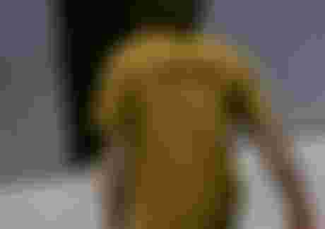 Man with long hair and a yellow T-shirt carrying surfboards towards a metal building with an open door.