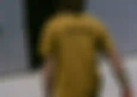 Man with long hair and a yellow T-shirt carrying surfboards towards a metal building with an open door.