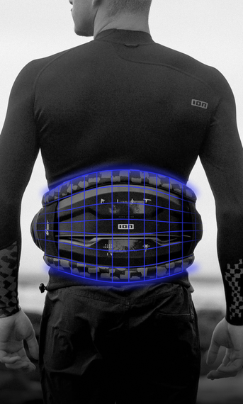 A person standing by the sea wearing a wetsuit and blue-highlighted harness, facing away with blurred water and rocks in the background.