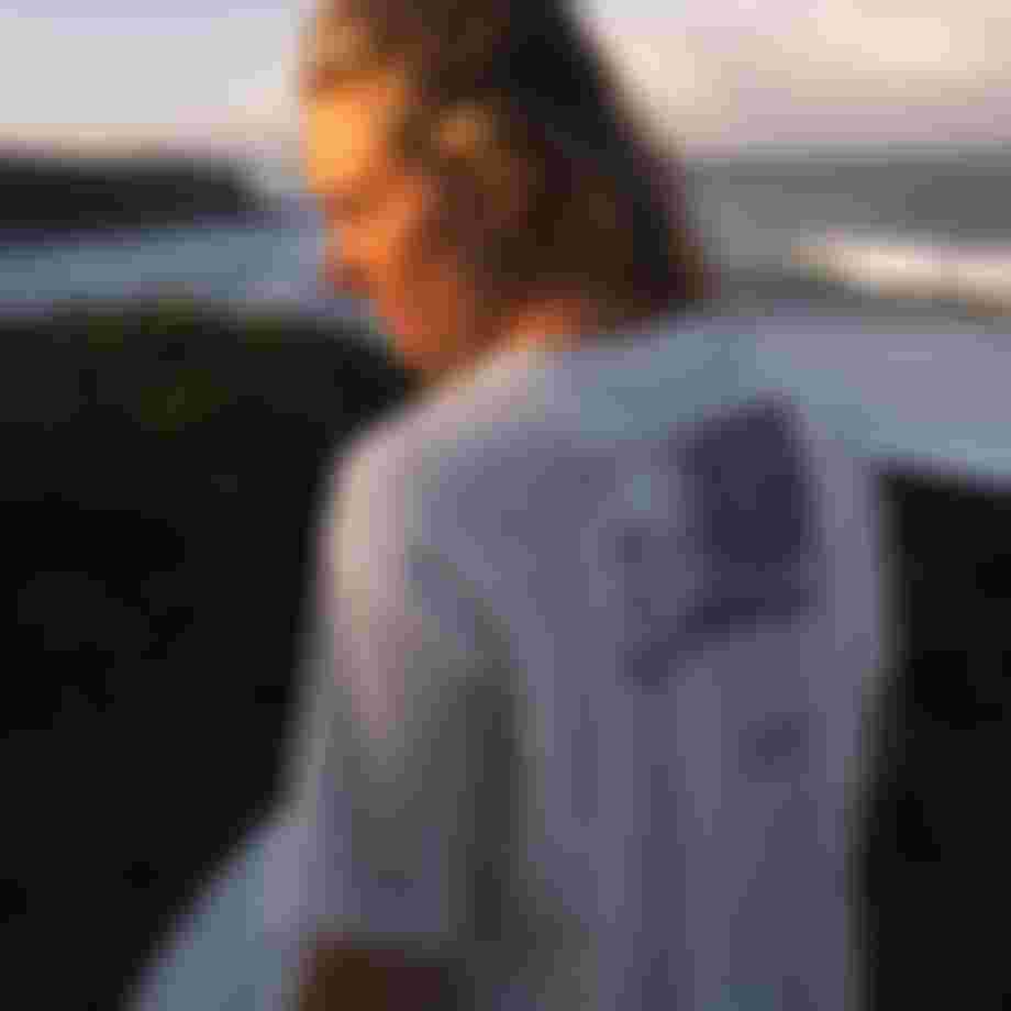 Person with sunlit hair faces the ocean, wearing a graphic white T-shirt with a purple design, against a backdrop of waves and cliffs.