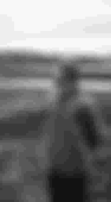 A person in a beanie and hoodie stands outdoors, gazing to the side, with a backdrop of hills and a cloudy sky. Black and white photo.