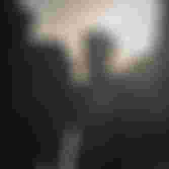 A man in a wetsuit stands on a rocky beach, backlit by the sun, surrounded by cliffs.