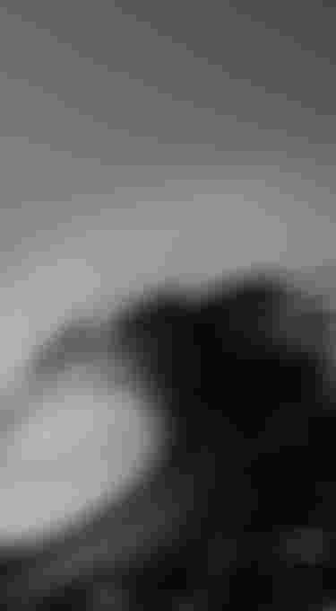 Black and white image of a large ocean wave curling, with water spray and a clear sky in the background.