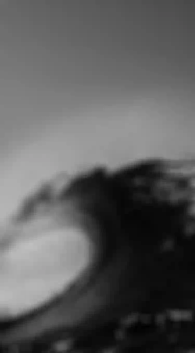 Black and white image of a large ocean wave curling, with water spray and a clear sky in the background.
