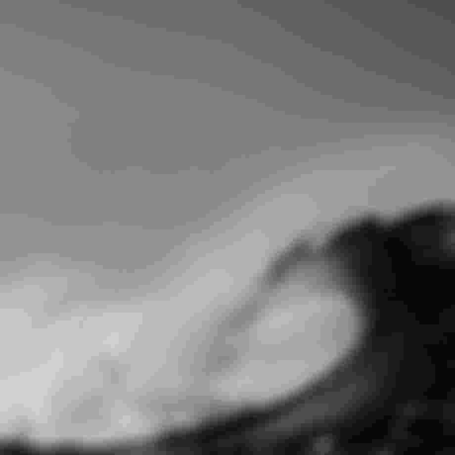 Black and white image of a large ocean wave curling, with water spray and a clear sky in the background.