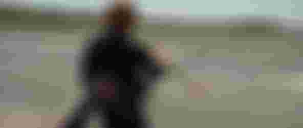 Person in wetsuit holding a kitesurfing board and kite lines, standing at the beach with gentle waves under a clear sky.