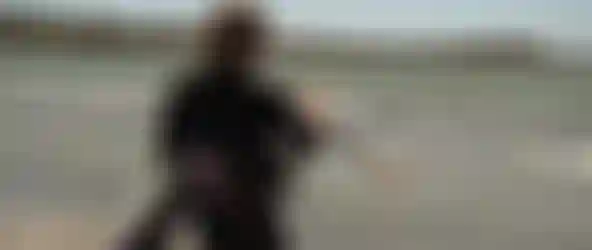 Person in wetsuit holding a kitesurfing board and kite lines, standing at the beach with gentle waves under a clear sky.