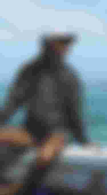 Woman in a gray hoodie and cap sits on a boat, gazing at the turquoise ocean under a clear blue sky.