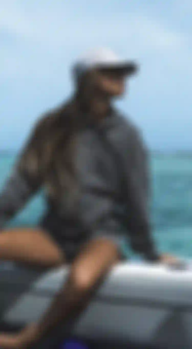 Woman in a gray hoodie and cap sits on a boat, gazing at the turquoise ocean under a clear blue sky.
