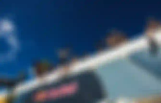 Spectators sitting on a Red Bull branded structure at an outdoor surf event under a deep blue sky, some holding surfboards.
