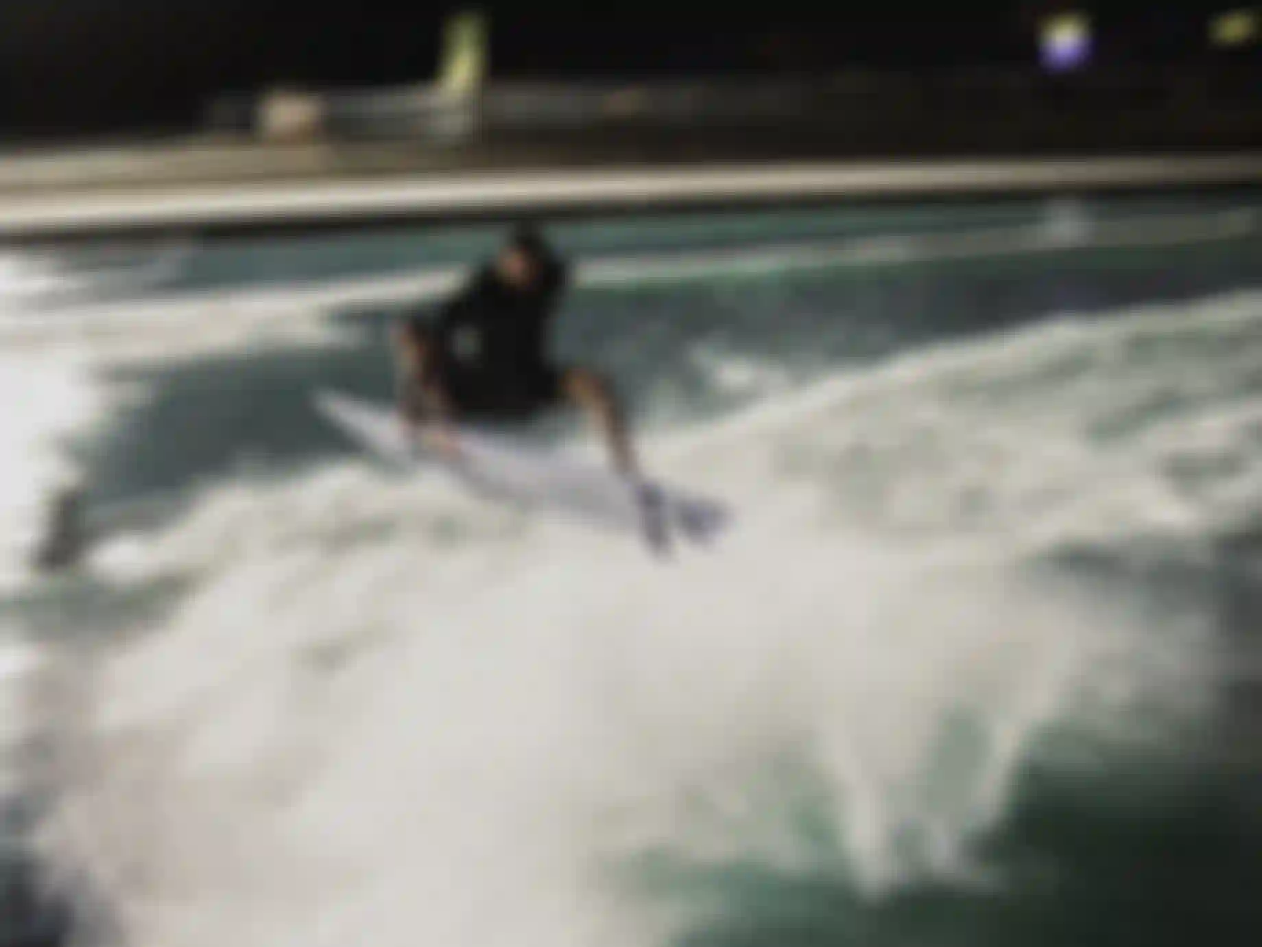 Surfer launching an aerial maneuver off a wave pool at night, board fully airborne above white water spray.