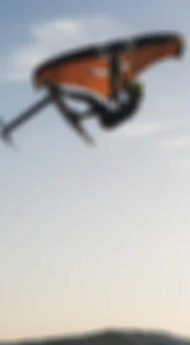 Person wing foiling in mid-air against a clear sky, performing an aerial maneuver with a bright orange wing and wearing a helmet.