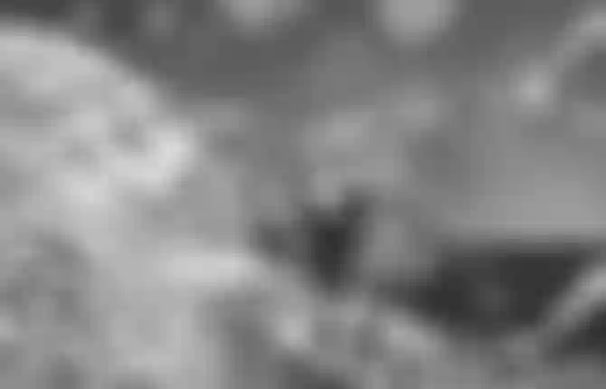 Black and white image of a person wearing a hat, kite surfing on a wave, with droplets on the lens and bright sunspots overhead.