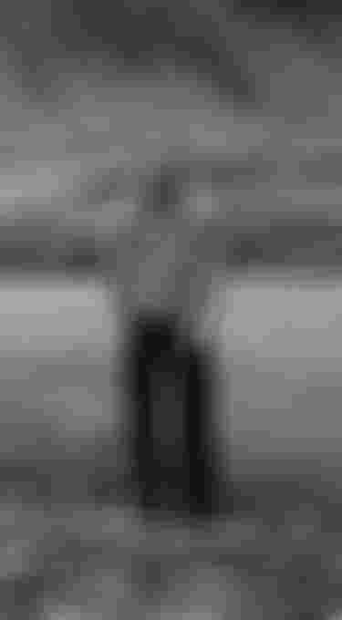 A person smiling and standing on a rocky riverbank, with hills in the background, captured in black and white.