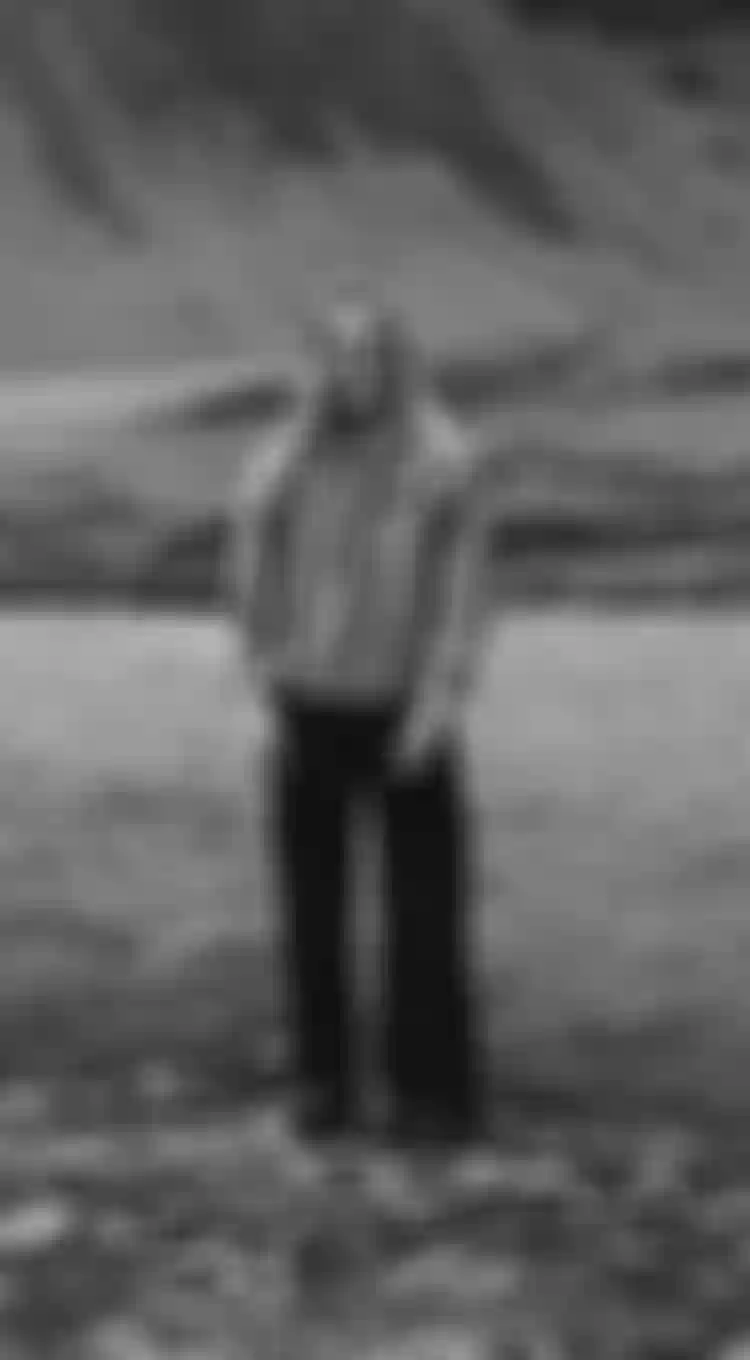 A person smiling and standing on a rocky riverbank, with hills in the background, captured in black and white.