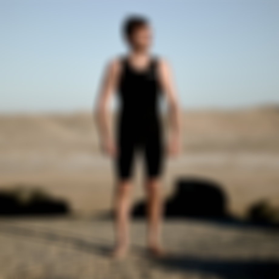 A person wearing a black wetsuit stands barefoot on a sandy, sunlit desert landscape, gazing into the distance.