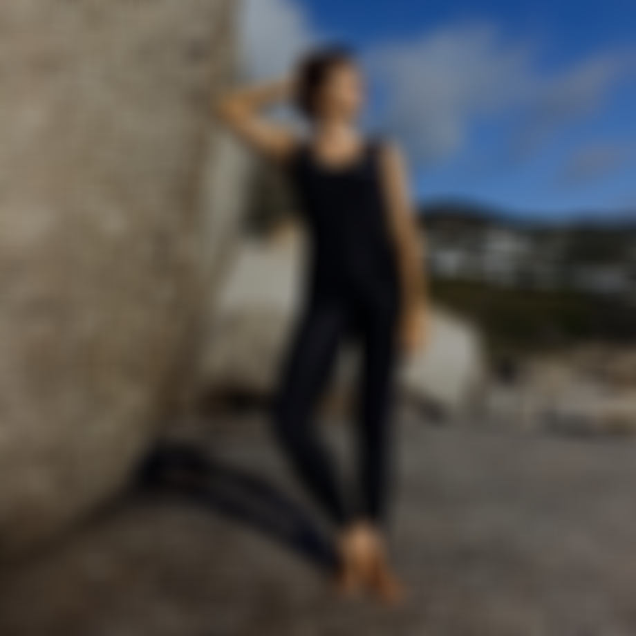 A woman in a black wetsuit stands barefoot on rocky terrain, leaning against a large boulder, with a coastal landscape in the background.