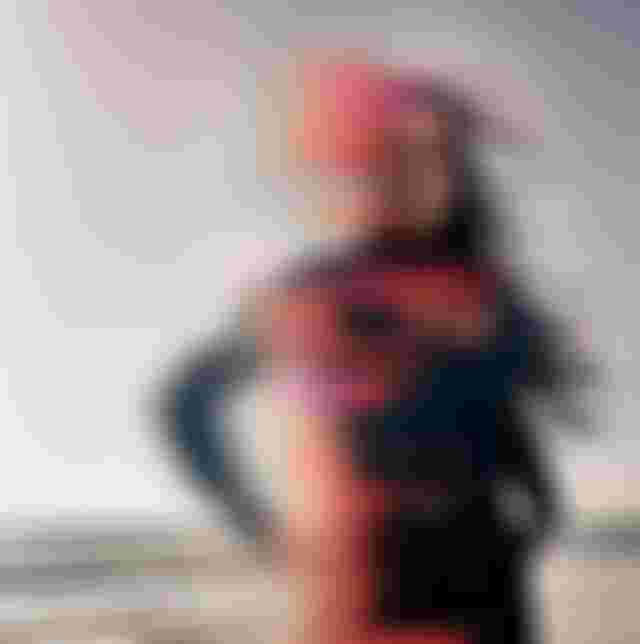 A woman in a wetsuit stands on a beach. Her hair is blown by the wind, with a colorful, glitch effect covering part of the image.