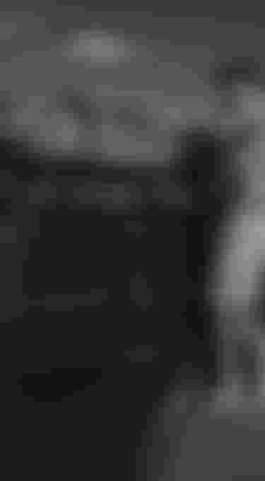 Man securing luggage on SUV roof near coastline, mountains in background, wearing cap and sunglasses.