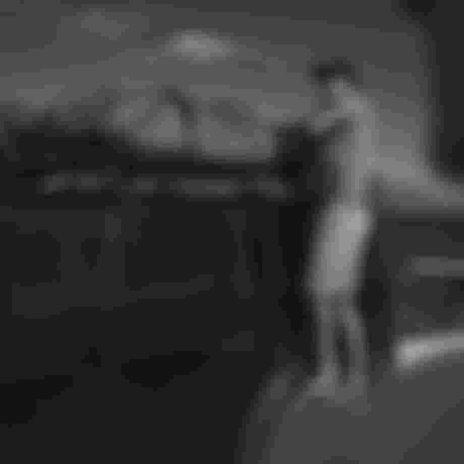 Man securing luggage on SUV roof near coastline, mountains in background, wearing cap and sunglasses.