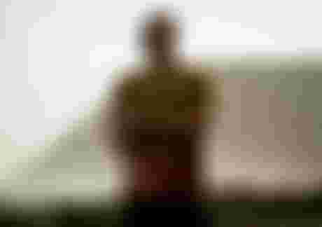 A smiling man in a brown T-shirt stands on a rocky beach with cliffs in the background, looking to the side in a relaxed pose.