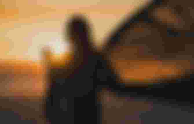 Silhouetted kite surfer holding gear at sunrise on a beach, with vibrant orange and yellow hues in the sky.