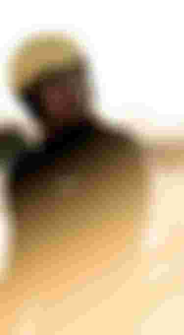 Person in a yellow helmet and wetsuit stands on a sandy beach with a surfboard, illuminated by soft sunlight.