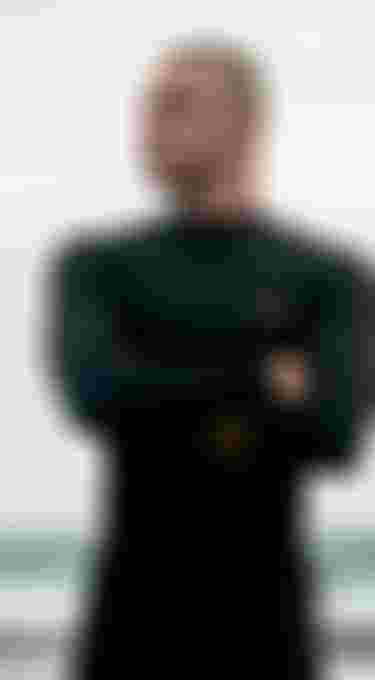 Man in black wetsuit with arms crossed stands on a beach, looking sideways. Waves and a cloudy sky in the background.